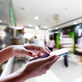 Una persona sosteniendo un teléfono celular mientras camina en un bullicioso centro comercial, rodeado de tiendas y gente.